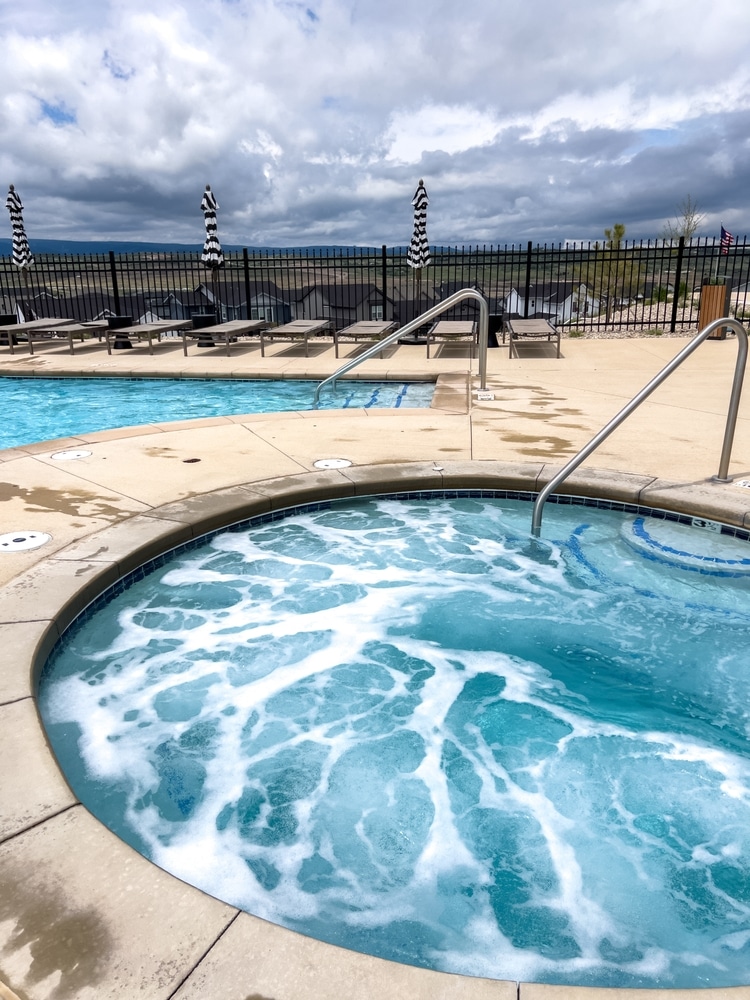 Bain à remous bouillonnant et piscine extérieure sous les nuages Bain à remous extérieur bouillonnant sur une terrasse de piscine. Chaises longues, parasols rayés et vue sur des maisons de banlieue sous un ciel très nuageux.
