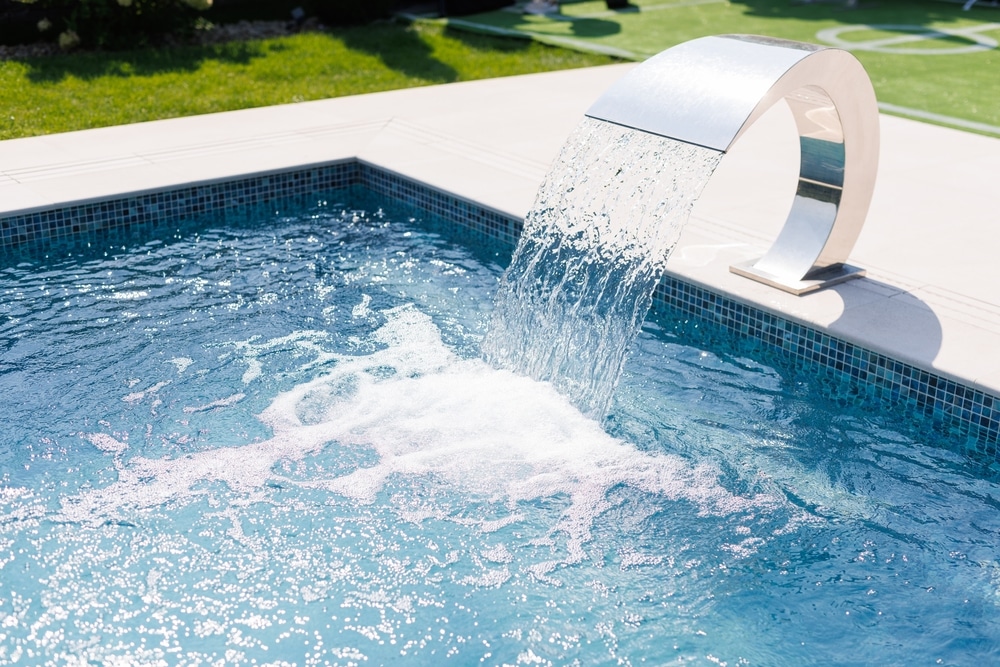 Cascade inox moderne pour piscine : ambiance luxe et relaxation. Luxueuse piscine extérieure avec cascade d'eau en acier inoxydable sur fond de mosaïque bleue. L'eau mousse sous le soleil.