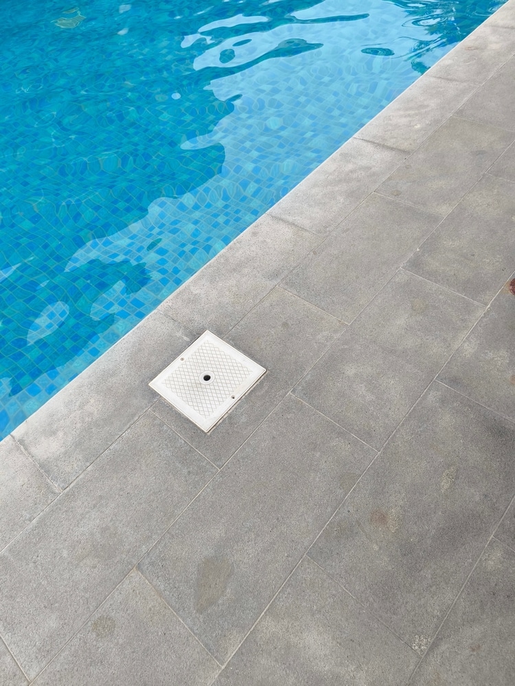 Dalles de piscine grises : la margelle moderne et épurée. Eau bleue agitée sur mosaïque de piscine, bordée de dalles grises. Couvercle de skimmer carré inséré dans la margelle.