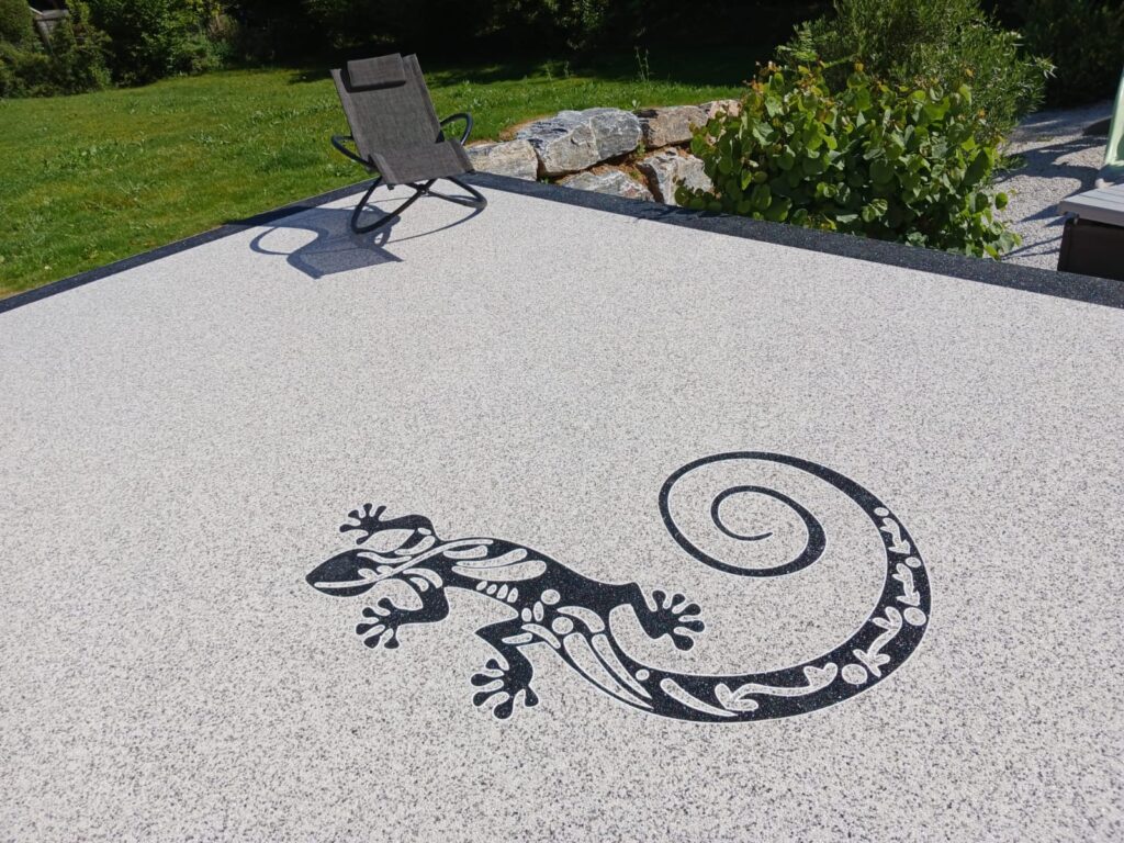 Gecko décoratif : Revêtement de sol de terrasse en résine. Terrasse claire en sol de résine moucheté décorée d'un grand gecko noir stylisé. Le transat et la verdure du jardin sont en arrière-plan.