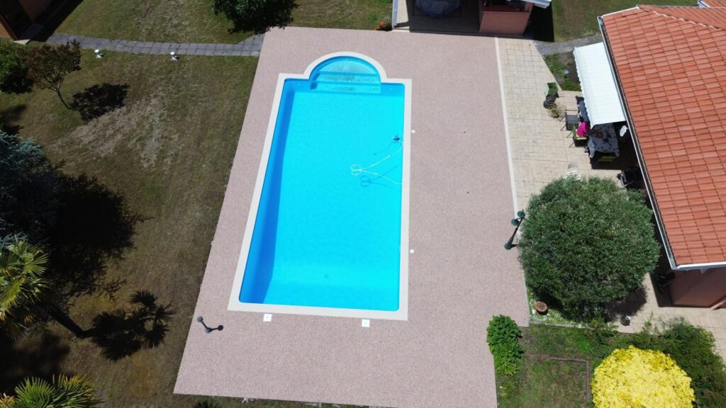 Piscine villa : design rectangulaire et terrasse en résine. Grande piscine extérieure d'un bleu vif avec escaliers, sur une terrasse en gravier de résine, avec jardin et villa en arrière-plan.