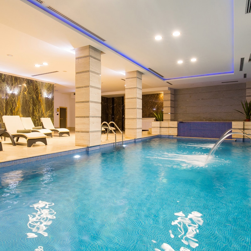 Piscine intérieure luxe : Détente au spa avec jet d'eau Piscine intérieure d'un spa de luxe. L'eau bleue reflète l'éclairage. Murs en marbre veiné, colonnes en pierre et transats pour la relaxation.