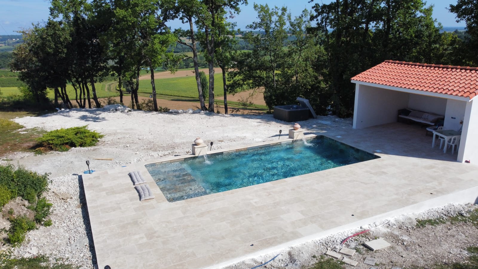 Piscine et terrasse en travertin : aménagement extérieur luxueux Piscine rectangulaire en travertin avec abri couvert et jacuzzi, aménagée sur un terrain en cours de finition, offrant une vue sur la campagne vallonnée.