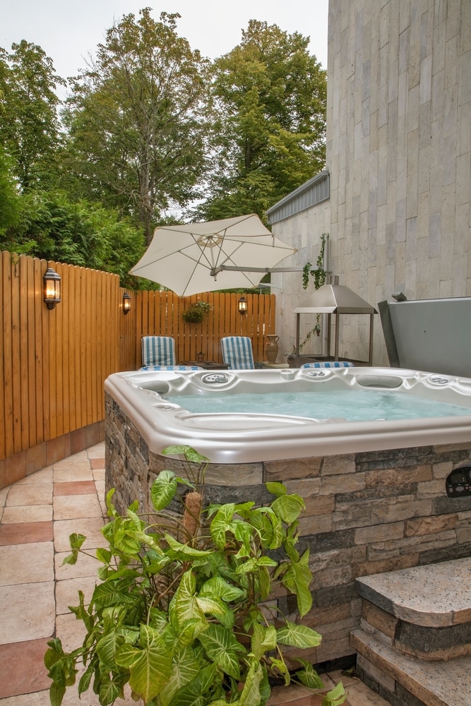 Jacuzzi extérieur sur patio privé : L'oasis de relaxation. Grand jacuzzi extérieur avec base en pierre et marches sur un patio. Clôture en bois, parasol blanc et végétation luxuriante.