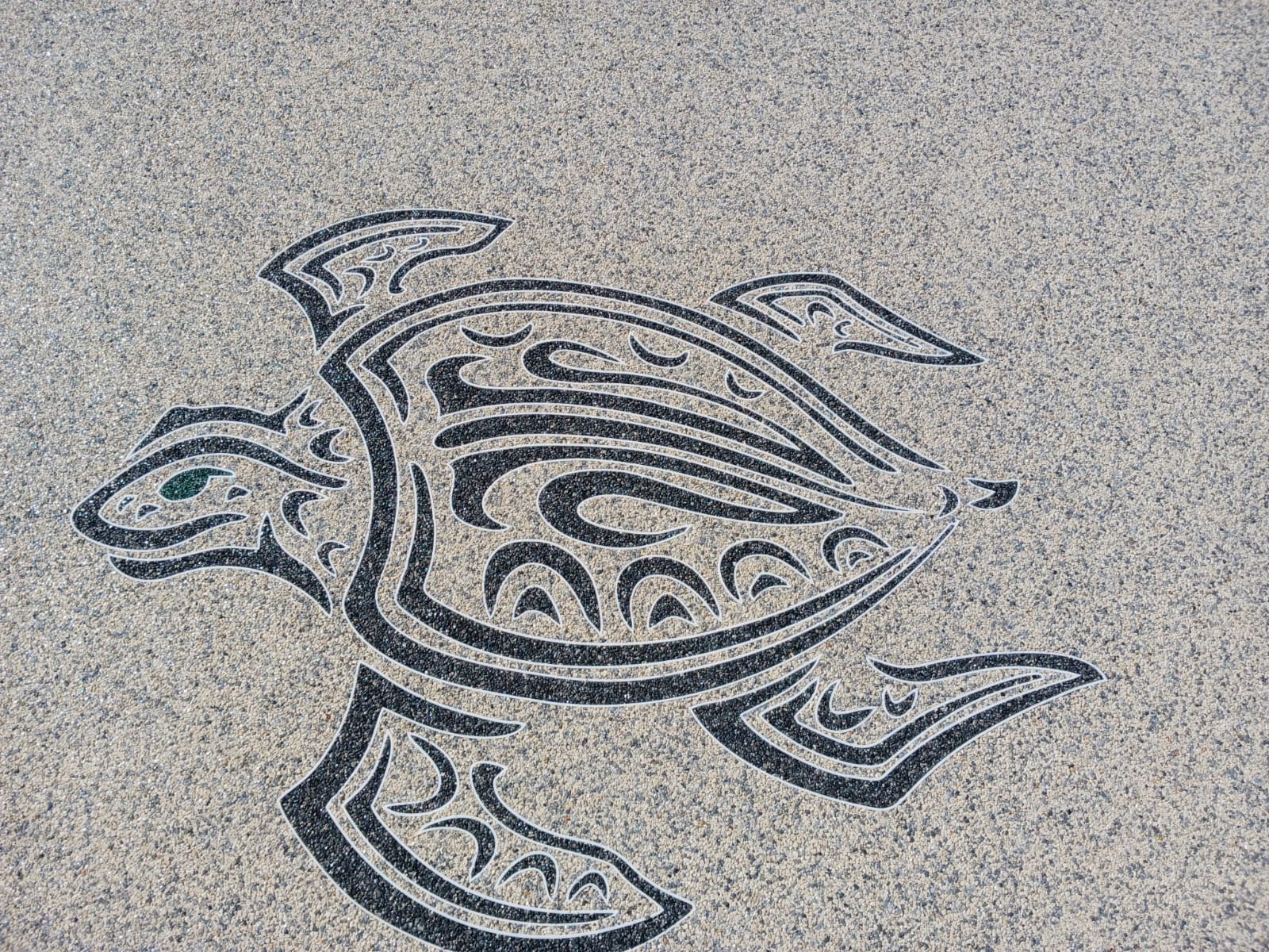 Tortue de mer stylisée (Honu) en motif tribal noir et blanc, gravée dans un sol texturé d'agrégats beiges. Œil vert.