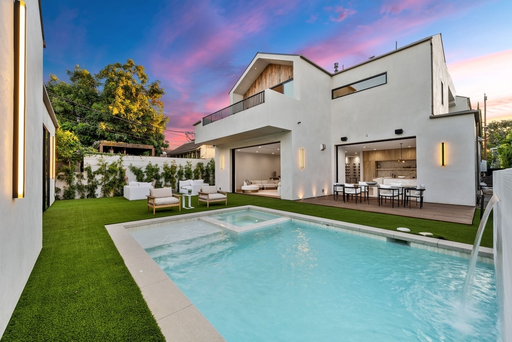 Maison moderne luxe : piscine, terrasse et coucher de soleil. Maison moderne blanche avec piscine, spa et terrasse en bois. La façade s'ouvre sur une cuisine intérieure au coucher du soleil rose.