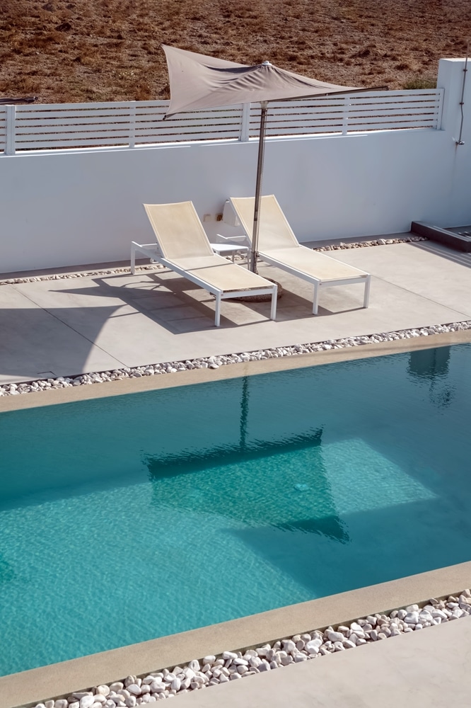 Piscine et transats : design minimaliste sous le soleil. Terrasse minimaliste et piscine turquoise ensoleillée. Deux transats blancs sous un parasol gris, près d'un mur blanc et de galets décoratifs.